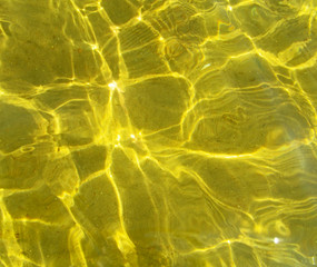 Sunlight refracting through transparent water and creates sun glare on golden sand of bottom. Top view of yellow ripple texture of liquid surface and sunny shining reflections.