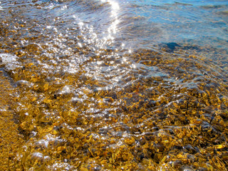 Transparent water and pebbles beach - close-up. Summer background image of sunlight reflecting and glistening on the water surface and wet small stones in foreground.