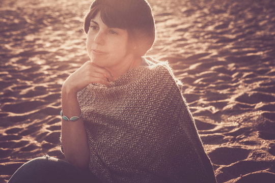 romantic portrait of middle age young caucasian girl woman sit down at the beach with sun and sunset in background. smile and emjoy the nature feeling looking ahead