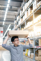 Young Asian man carrying cardboard box over head between row of shelves in warehouse, shopping warehousing or working pick and packing concepts