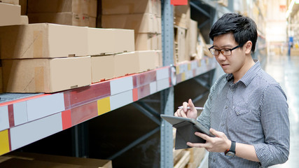 Young Asian man worker doing stocktaking of product in cardboard box on shelves in warehouse by using digital tablet and pen. Physical inventory count concept