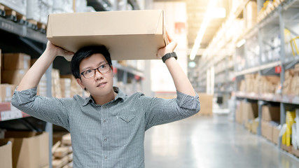 Young Asian man carrying cardboard box over head between row of shelves in warehouse, shopping warehousing or working pick and packing concepts