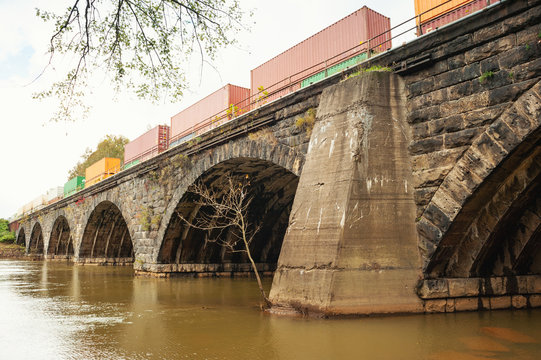 A skewed arch stone bridge in Western Pennsylvania stretching across a river.  A cargo train is passing over the bridge.