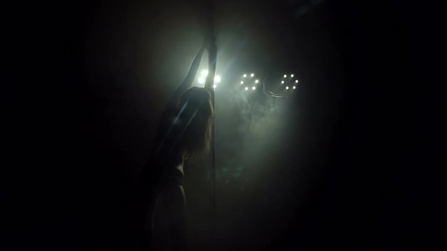 Silhouette of a girl with long flowing hair dancing on a pole in a dark smoke-filled strip club. Slow motion. Pole dance.