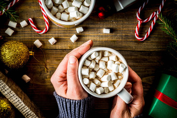 Girl drinks hot chocolate with marshmallow, cocoa mug in woman hand in warm winter sweater, old wooden table  with christmas decorations top view copy space