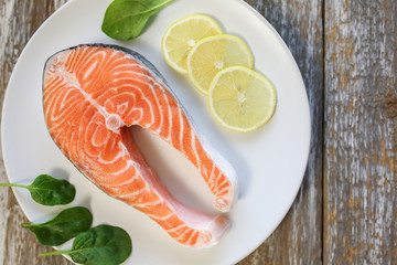 A piece of fresh salmon fish steak on a white plate with spinach and lemon, on  a gray wooden background. Omega 3 vitamin, healthy lifestyle. Natural vegetarian food. Top view.