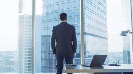 Back View of the Thoughtful Businessman wearing a Suit Standing in His Office, Hands in Pockets and Contemplating Next Big Business Deal, Looking out of the Window. Big City Panoramic Window View. - obrazy, fototapety, plakaty