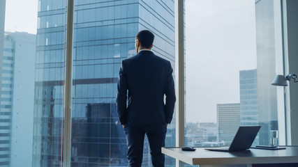 Back View of the Thoughtful Businessman wearing a Suit Standing in His Office, Hands in Pockets and Contemplating Next Big Business Deal, Looking out of the Window. Big City Panoramic Window View. - obrazy, fototapety, plakaty