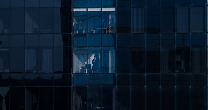 Aerial View Shot: From Outside into Office Building with Businessman Using Mobile Phone and Standing Next to the Office Window. Beautiful Shot of The Financial Business District Skyscrapers. 