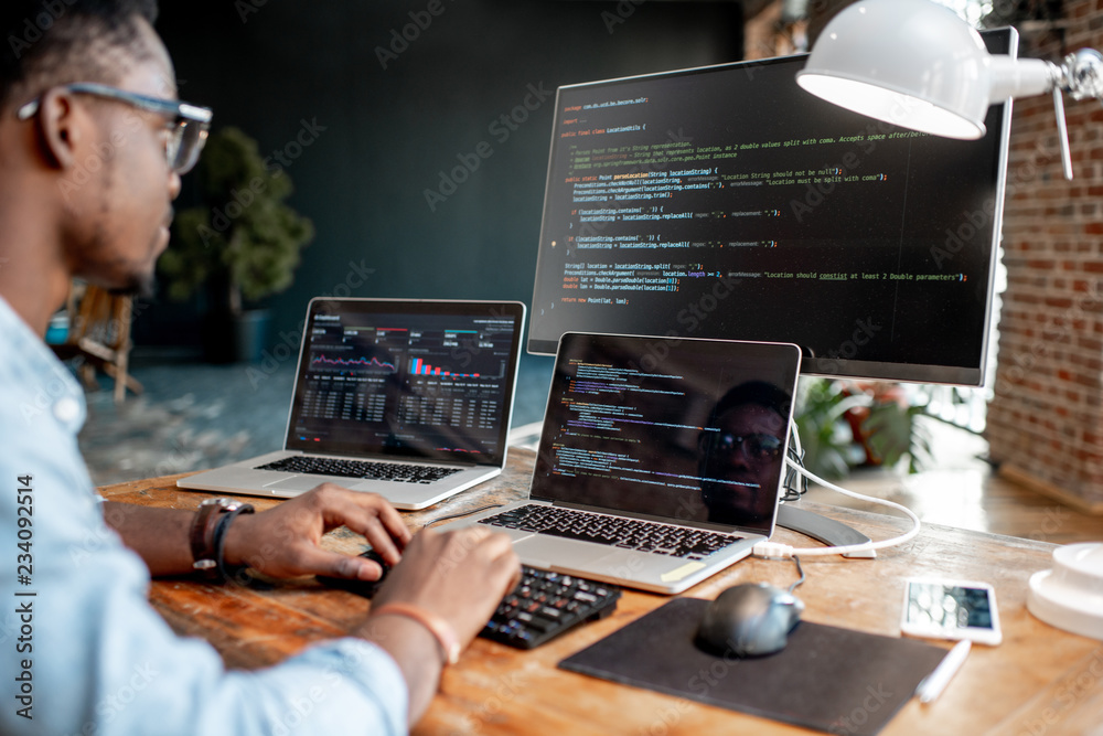 Wall mural young african male programmer writing program code sitting at the workplace with three monitors in t