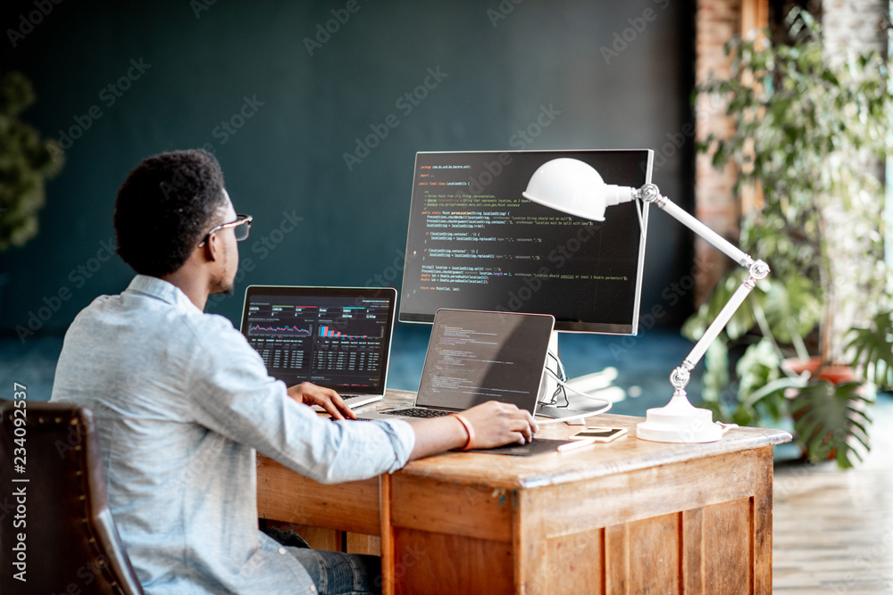 Wall mural young african male programmer writing program code sitting at the workplace with three monitors in t