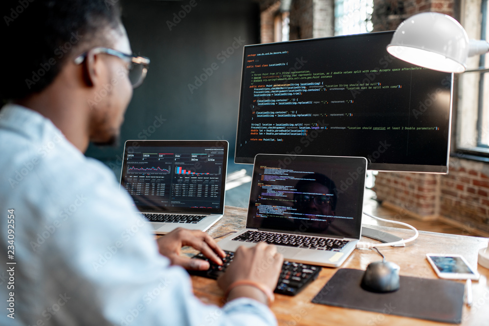Wall mural young african male programmer writing program code sitting at the workplace with three monitors in t