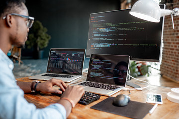 Young african male programmer writing program code sitting at the workplace with three monitors in the office. Image focused on the screen - obrazy, fototapety, plakaty