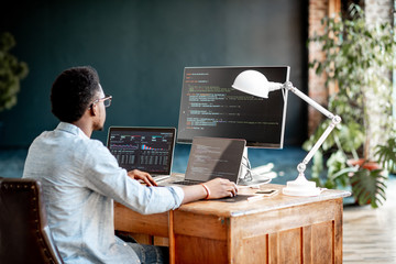 Young african male programmer writing program code sitting at the workplace with three monitors in...