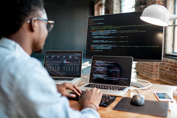 Young african male programmer writing program code sitting at the workplace with three monitors in the office. Image focused on the screen - Powered by Adobe