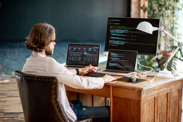 Young male programmer writing a program code sitting at the workplace with three monitors in the...