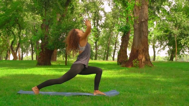 caucasian sportswoman practicing yoga at the open air slow motion girl with long curly hair trains outdoors. nature landscape with green grass and trees