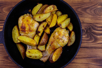 Baked chicken drumsticks with potatoes in in cast-iron frying pan on wooden table. Top view