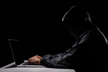 A male hacker cover his head with the hood trying to steal the data from a laptop computer, dark...