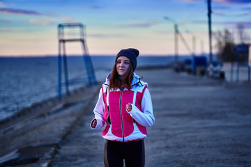 A young girl in a sports hat and jacket makes a morning jog on the embankment in the morning before...