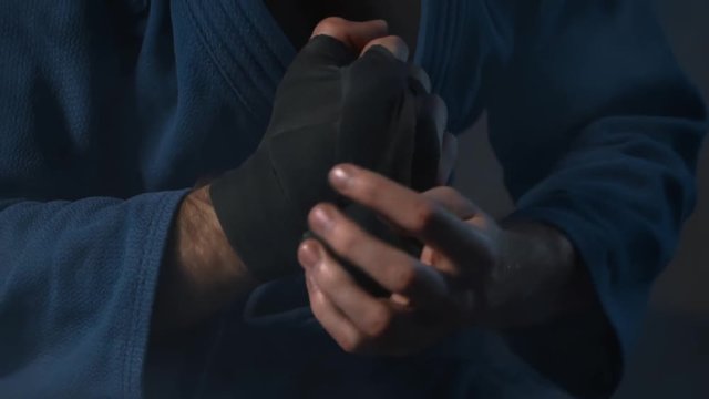 MMA Fighter Preparing For Training. Closeup View Male Wrapping Bandage On Hand. Adult wrestler dressed in blue kimono prepare fist for workout indoor in gym. Professional Athlete ready to fight