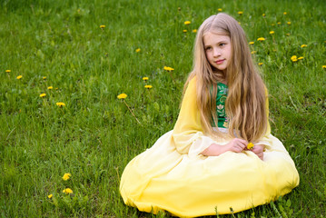 Blond young girl posing in a yellow green dress sitting on the grass with dandelions yellow flowers