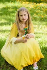 Blond young girl posing in a yellow green dress sitting on the grass with dandelions yellow flowers