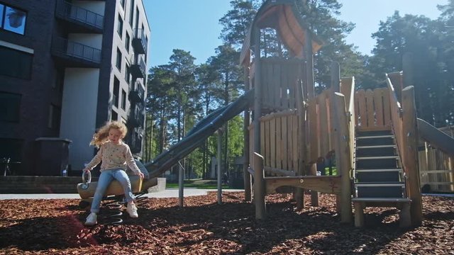 Happiness moments, family concept. Sweet girl play at public playground at sunny summer day near with residental apartaments or hotel. Casual daily joyful activity, at background green forest or park