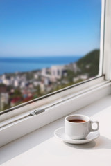 white cup with drink on a window sill with blurred city view with  blue sky and sea copy space