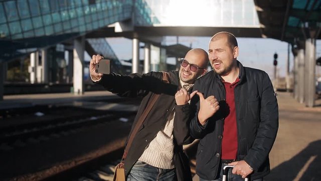 Two men on the platform of the railway station waiting for the arrival of the train and take pictures selfie on a smartphone.