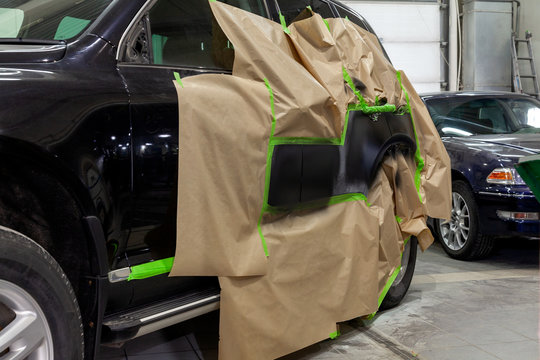 Partial Painting Of The Side Elements Of The Body Of A Black SUV Car After An Accident On The Road, Not Damaged Parts Are Covered With Paper And Tape From Getting Into Paint In A Car Repair Workshop