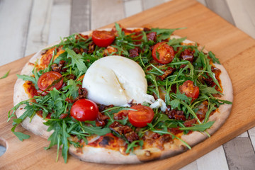 A pizza on a wooden board with cherry tomatoes, arugula, and burrata creamy cheese