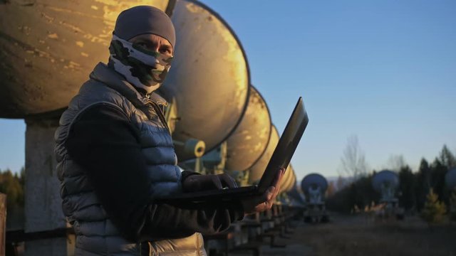 Hacker hacks the radio telescope using notebook. Man to program hacking on laptop in nature. Terrorist is hiding in camouflage balaclava. Hacker has criminal cases on the Internet. Programmer uses a