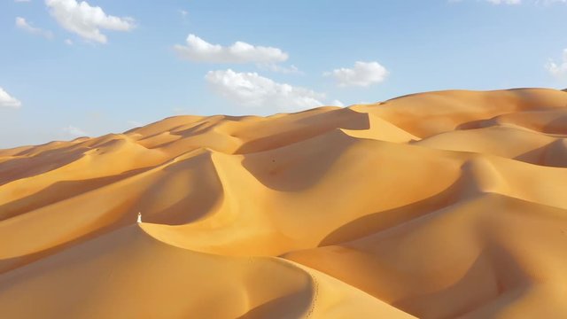 Aerial drone view of a young woman in white summer dress and panama hat standing on top of a massive sand dune with her skirt flying in the wind. Abu Dhabi, UAE.