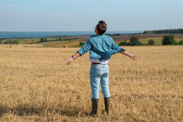 Naklejka na ściany i meble Young man in jeans, shirt, rubber boots in the field with his hands open, the concept of freedom, motivation, movement, portrait from the back