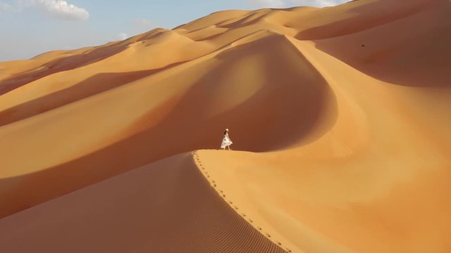 Young woman in a white summer dress walking on top of a massive sand dune. Empty Quarter, Abu Dhabi, UAE.
