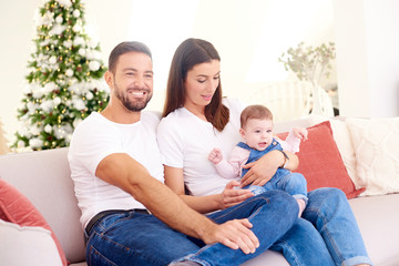 Happy young parents with their baby girl enjoy Christmas time while relaxing on the sofa. Family life.