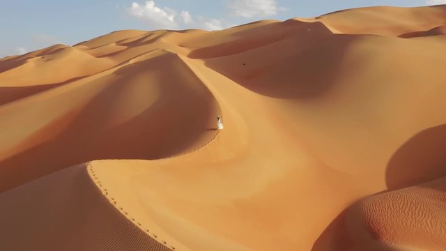 Aerial drone view of a young woman in white summer dress and panama hat walking up massive yellow and gold sand dunes with shadows in empty quarter during sunset. Abu Dhabi, UAE.