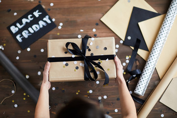 Directly above view of female hands holding wrapped gift box with ribbon bow and confetti over wooden table with card Black Friday