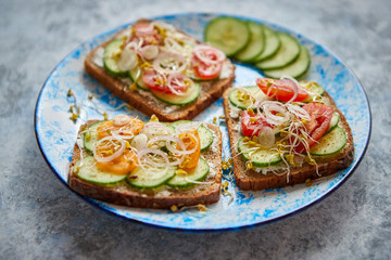 Plate with healthy toasts with cucomber, cherry tomatoes, crumbled feta cheese and radish sprouts on a black plate. Table top view. Gray stone background.