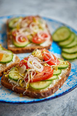 Plate with healthy toasts with cucomber, cherry tomatoes, crumbled feta cheese and radish sprouts on a black plate. Table top view. Gray stone background.
