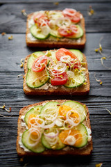 Row of three healthy toasts with onion, cucomber, cherry tomatoes, crumbled feta cheese and radish sprouts on a black plate. Table top view on black wooden background