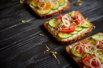 Row of three healthy toasts with onion, cucomber, cherry tomatoes, crumbled feta cheese and radish sprouts on a black plate. Table top view on black wooden background