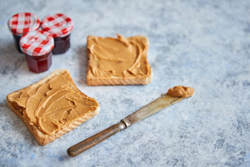 Fototapeta na wymiar Two tasty peanut butter toasts placed on stone table. With small jars of fruit jam. Knife on side. Above view.