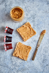 Two tasty peanut butter toasts placed on stone table. With small jars of fruit jam big peanut butter container. Knife on side. Above view.