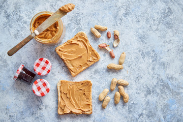 Two tasty peanut butter toasts placed on stone table. With small jars of fruit jam big peanut butter container. Knife on side. Above view with copy space