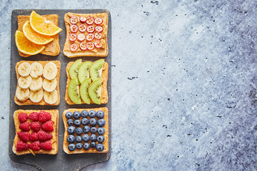 Six Healthy breakfast toasts. Wholegrain bread slices with peanut butter and various fruits. Served on grey cutting board. Top view, grey stone background. Dieting concept with cpoy space