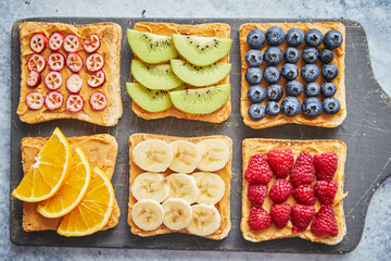 Healthy breakfast toasts. Wholegrain bread slices with peanut butter and various fruits. Served on grey cutting board. Top view, grey stone background. Dieting concept