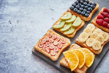 Six Healthy breakfast toasts. Wholegrain bread slices with peanut butter and various fruits. Served on grey cutting board. Top view, grey stone background. Dieting concept with cpoy space