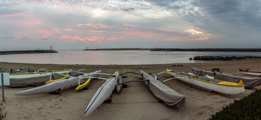 Panoramic scene of golden hour clouds over outrigger rower boats of Ventura Harbor Cove under Autumn dusk sky. © motionshooter